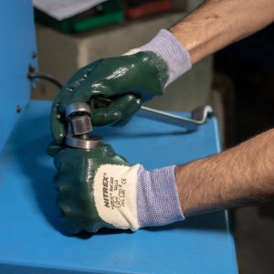 Operario usando guantes de seguridad NITREX 220 mientras ajusta una pieza metálica en maquinaria industrial azul.