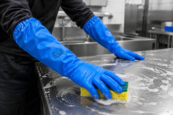 Operario utilizando guantes largos NITREX 314LNIT azules durante la limpieza de una superficie industrial con espuma y productos de limpieza.