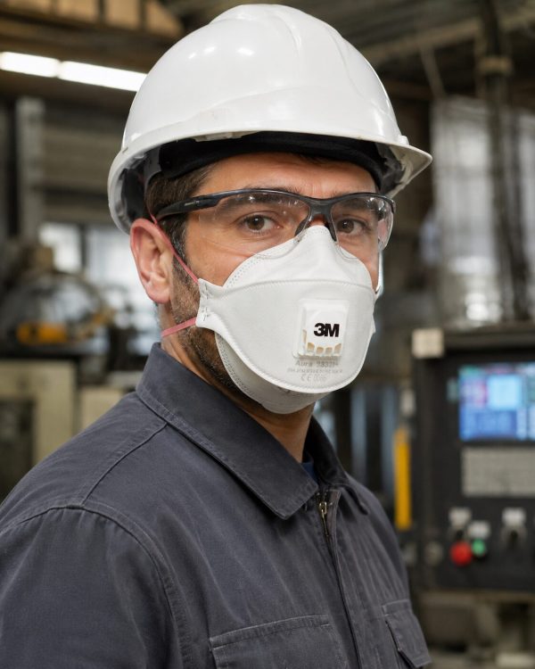 Operario con casco y gafas de seguridad utilizando mascarilla 3M Aura 9332 Gen3 FFP2 con válvula en fábrica industrial