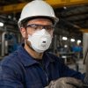 Trabajador industrial con casco y gafas de seguridad usando mascarilla 3M Aura 9332+ FFP3 con válvula en entorno de fábrica.