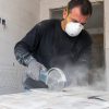 Operario de la construcción con guantes y mascarilla 3M 8710 FFP1 cortando un azulejo con radial en un ambiente con polvo en suspensión.