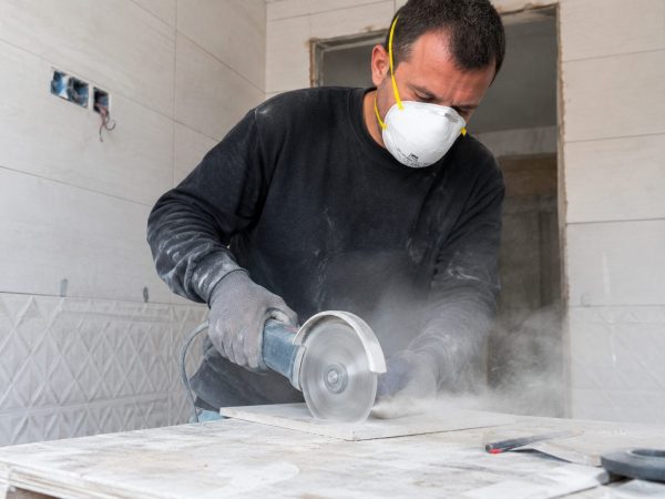 Operario de la construcción con guantes y mascarilla 3M 8710 FFP1 cortando un azulejo con radial en un ambiente con polvo en suspensión.