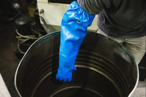 Trabajador usando guante largo impermeable NITREX 314LNIT azul para manipular líquidos y recipientes en entorno industrial.