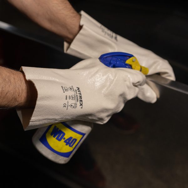 Operario con guantes de nitrilo blanco Nitrex 420BL aplicando espray lubricante WD-40 en una superficie metálica.